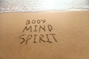 The words "Body Mind Spirit" are written in the sand near the edge of a wave on a beach.